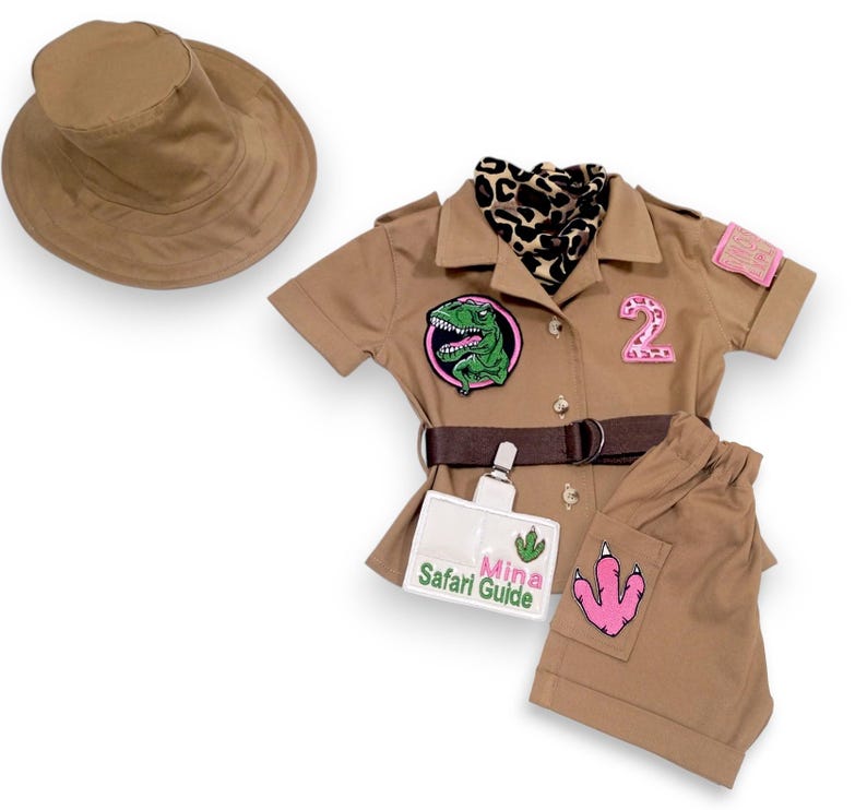 Puede incluir: Un conjunto de disfraz de safari color canela para ni&ntilde;os, que incluye una camisa con botones, pantalones cortos y un sombrero de ala ancha. La camisa presenta un parche de dinosaurio, un parche con el n&uacute;mero 2 y un cintur&oacute;n marr&oacute;n. Los pantalones cortos tienen un parche rosa con una huella de dinosaurio.