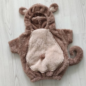 Puede incluir: Un disfraz de mono de peluche marr&oacute;n con capucha y cola. El disfraz tiene una barriga marr&oacute;n claro y una cara marr&oacute;n.