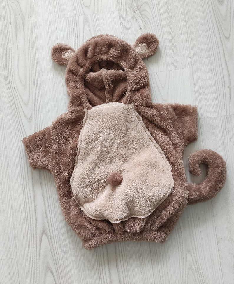 Puede incluir: Un disfraz de mono de peluche marr&oacute;n con capucha y cola. El disfraz tiene una barriga marr&oacute;n claro y una cara marr&oacute;n.
