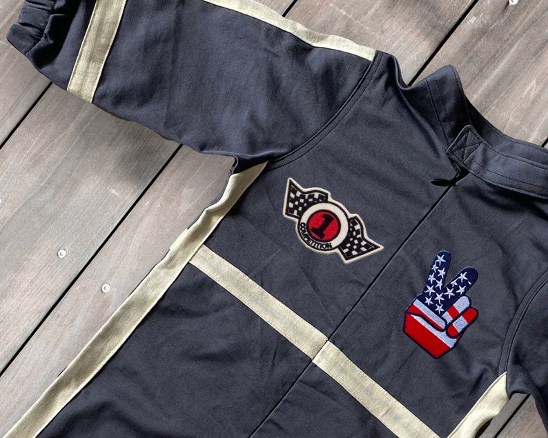 May include: A black jumpsuit with white stripes and two patches. One patch is a red circle with the number "1" and the words "Competition" around it. The other patch is a red, white, and blue peace sign.