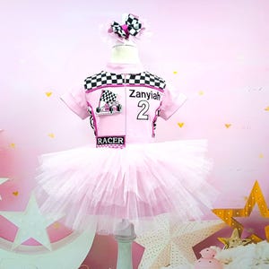 May include: Pink tutu dress with a black and white checkered racing top. The top has the name "Zaniyah" and the number "2" on it. The top also has a pink and black race car graphic. The dress is on a white stand against a pink background.