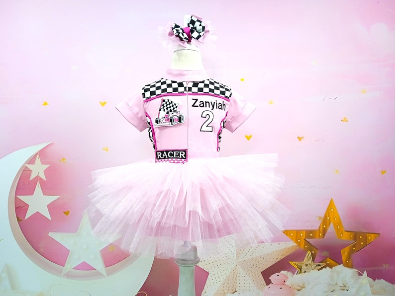 May include: Pink tutu dress with a black and white checkered racing top. The top has the name "Zaniyah" and the number "2" on it. The top also has a pink and black race car graphic. The dress is on a white stand against a pink background.