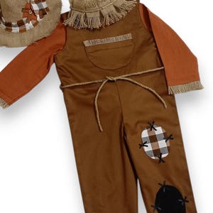 May include: A brown scarecrow costume featuring long sleeves, a burlap-style collar, and a matching hat. The overalls include a chest pocket, a rope tie at the waist, and two patches on the leg: one brown and white plaid, one solid black.