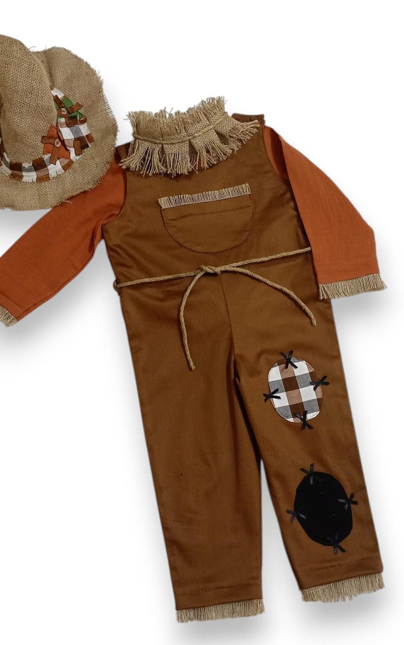 May include: A brown scarecrow costume featuring long sleeves, a burlap-style collar, and a matching hat. The overalls include a chest pocket, a rope tie at the waist, and two patches on the leg: one brown and white plaid, one solid black.
