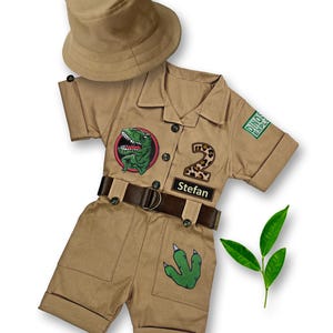 May include: A tan safari-themed jumpsuit with a dinosaur graphic, a leopard print number 2, and a name tag that says "Stefan". The jumpsuit has a brown belt and a green dinosaur footprint on the leg. A tan safari hat is included.