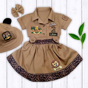 May include: A brown safari-themed dress with a leopard print trim, a matching hat with ears, and a bow. The dress has patches that say "Safari" and "Lillian" and a number "2" patch.