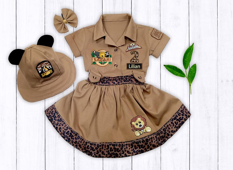 May include: A brown safari-themed dress with a leopard print trim, a matching hat with ears, and a bow. The dress has patches that say "Safari" and "Lillian" and a number "2" patch.