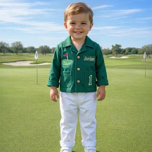 May include: A young child wearing a green golf jacket with gold buttons, a golf flag patch, and the number 1. The jacket has the name "Jordan" on it. The child is also wearing white pants and shoes, standing on a green golf course.