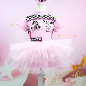 May include: Pink tutu dress with a black and white checkered racing top. The top has a pink racing car graphic and the name "Zanyiah" with the number "2" on it. The top also has the word "RACER" printed on it.
