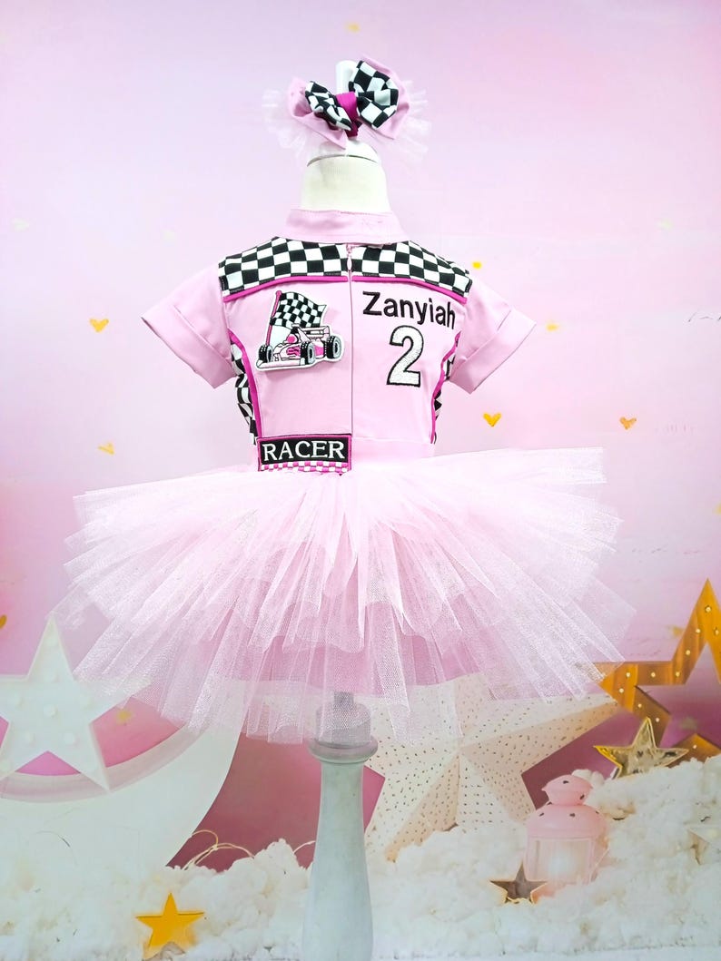 May include: Pink tutu dress with a black and white checkered racing top. The top has a pink racing car graphic and the name "Zanyiah" with the number "2" on it. The top also has the word "RACER" printed on it.
