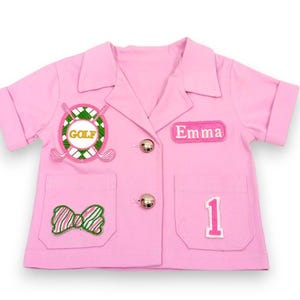 May include: A pink short-sleeved jacket with gold buttons and pockets. The jacket has an embroidered "GOLF" emblem, a bow, the name "Emma", and the number "1". The jacket is designed for a child.