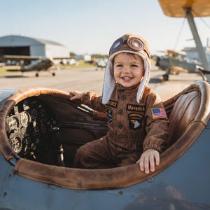 May include: A brown flight suit with patches reading Air Force, Maverick, and Airborne, featuring an American flag patch on the sleeve. The outfit includes a brown aviator cap with white faux fur lining and brown goggles resting on top.