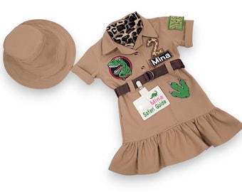 Puede incluir: Un vestido de safari de color marrón claro con un parche de dinosaurio, una etiqueta de nombre que dice "Mina Safari Guide" y un cinturón marrón. El vestido tiene un dobladillo con volantes y mangas cortas. También se incluye un sombrero marrón claro.