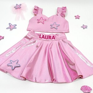 May include: Pink two-piece outfit for girls with a sparkly skirt and a crop top. The skirt has a pink elastic waistband with the name "LAURA" printed on it. The crop top has a sweetheart neckline and is decorated with pink and purple starfish.