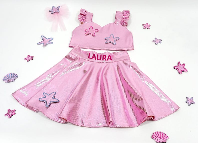 May include: Pink two-piece outfit for girls with a sparkly skirt and a crop top. The skirt has a pink elastic waistband with the name "LAURA" printed on it. The crop top has a sweetheart neckline and is decorated with pink and purple starfish.