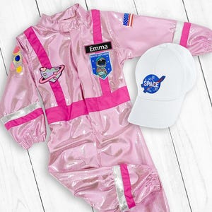 May include: Pink astronaut costume with a white hat that says "SPACE" on it. The costume has a name tag that says "Emma" and patches of a rocket, a planet, and an astronaut.