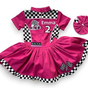May include: A hot pink racing-themed outfit with a matching headband. The outfit features a collared shirt with a checkered pattern, the name "Emma" and the number "2". The skirt has a full, pleated design with checkered trim and racing flags.