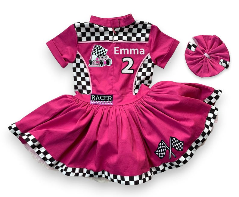 May include: A hot pink racing-themed outfit with a matching headband. The outfit features a collared shirt with a checkered pattern, the name "Emma" and the number "2". The skirt has a full, pleated design with checkered trim and racing flags.