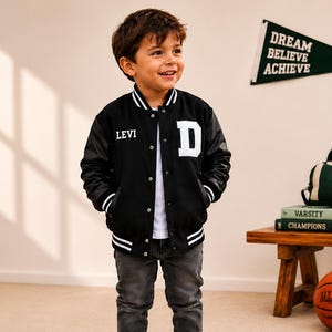 May include: A black varsity jacket with long black sleeves, white striped ribbed cuffs, a white striped collar, and a white snap-button front. The jacket features the name LEVI in white text on the right chest and a large white letter D on the left.