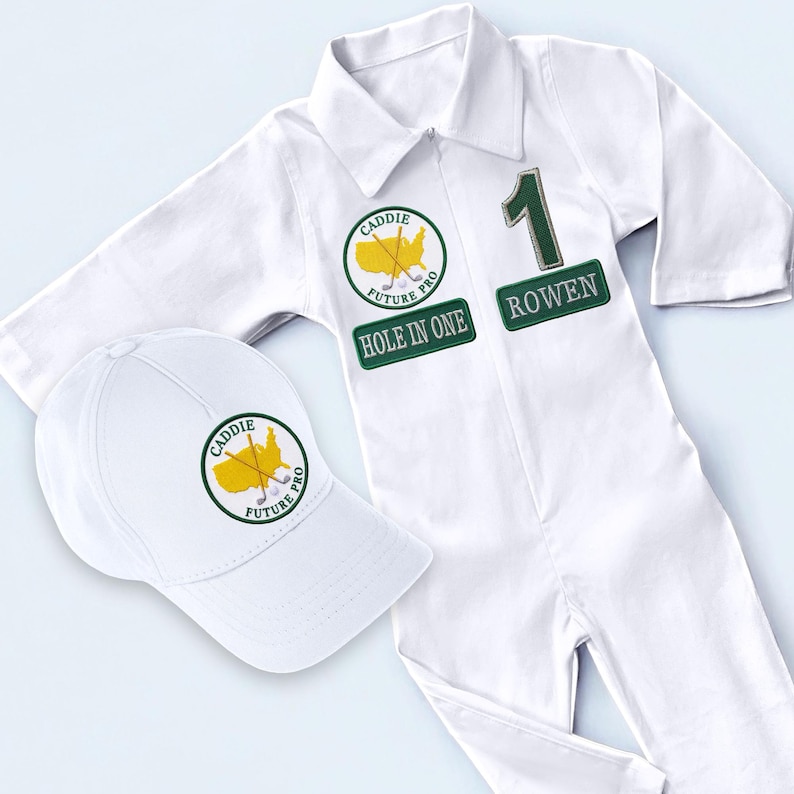 A high-quality white cotton golf caddy jumpsuit for kids featuring professional green embroidery with the name Rowen and a number 1 patch. The outfit includes a matching white baseball cap with a circular golf logo, all displayed on a clean white background. This set is designed to mimic a professional Masters-style tournament uniform.