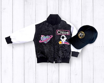 Puede incluir: Chaqueta bomber negra y blanca con lentejuelas y parches coloridos, incluyendo un corazón con un planeta, un astronauta y una etiqueta con el nombre "Chloe". También se incluye una gorra de béisbol negra con un diseño bordado en dorado.