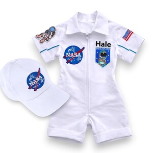 May include: A white NASA-themed jumpsuit and matching baseball cap. The jumpsuit features a zipper, blue accents, and patches with the NASA logo, an astronaut, and the text "Hale SPACE EXPLORER". The cap also displays the NASA logo.