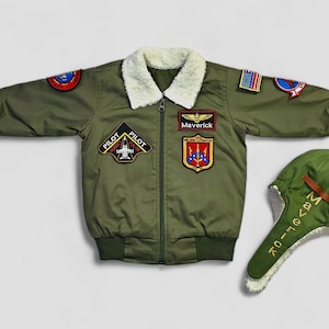 K&ouml;nnte beinhalten: Eine olivgr&uuml;ne Bomberjacke mit wei&szlig;em Kunstfellkragen, Frontrei&szlig;verschluss und verschiedenen gestickten Aufn&auml;hern, darunter ein Maverick-Namensschild, ein Pilot Hornet F/A-18-Emblem und eine US-Flagge. Enth&auml;lt eine passende olivgr&uuml;ne Fliegerm&uuml;tze.
