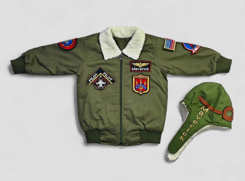 K&ouml;nnte beinhalten: Eine olivgr&uuml;ne Bomberjacke mit wei&szlig;em Kunstfellkragen, Frontrei&szlig;verschluss und verschiedenen gestickten Aufn&auml;hern, darunter ein Maverick-Namensschild, ein Pilot Hornet F/A-18-Emblem und eine US-Flagge. Enth&auml;lt eine passende olivgr&uuml;ne Fliegerm&uuml;tze.