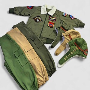K&ouml;nnte beinhalten: Eine olivgr&uuml;ne Bomberjacke mit wei&szlig;em Kunstfellkragen, verziert mit verschiedenen Luftfahrt-Aufn&auml;hern und einem Maverick-Namensschild. Dazu geh&ouml;ren zwei Cargohosen in Oliv und Beige sowie zwei Fliegerm&uuml;tzen mit Kunstfellfutter.