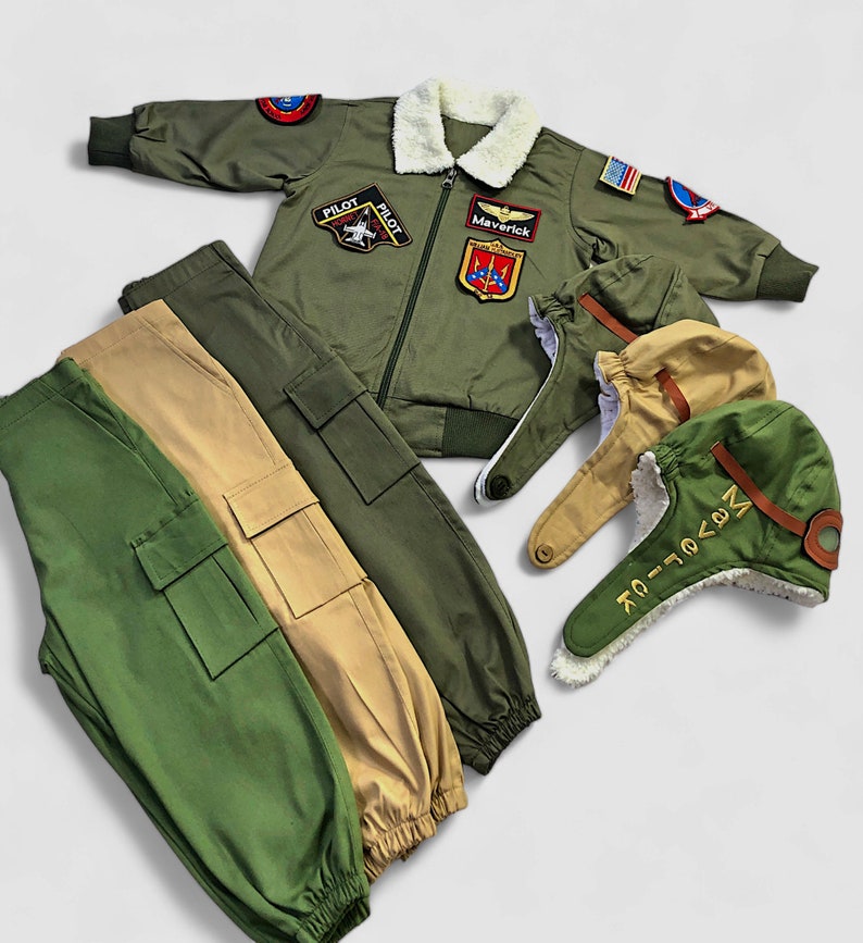 K&ouml;nnte beinhalten: Eine olivgr&uuml;ne Bomberjacke mit wei&szlig;em Kunstfellkragen, verziert mit verschiedenen Luftfahrt-Aufn&auml;hern und einem Maverick-Namensschild. Dazu geh&ouml;ren zwei Cargohosen in Oliv und Beige sowie zwei Fliegerm&uuml;tzen mit Kunstfellfutter.