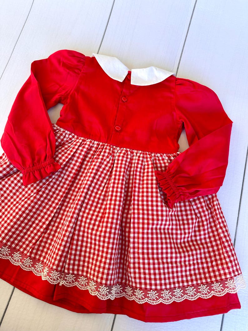 K&ouml;nnte beinhalten: Rot-wei&szlig;es Gingham-Kleid mit wei&szlig;em Peter-Pan-Kragen und langen &Auml;rmeln. Das Kleid hat einen Spitzenbesatz am Saum.