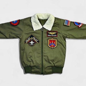 K&ouml;nnte beinhalten: Eine olivgr&uuml;ne Bomberjacke mit wei&szlig;em Kunstfellkragen, Frontrei&szlig;verschluss und gerippten B&uuml;ndchen. Die Jacke ist mit verschiedenen gestickten Aufn&auml;hern verziert, darunter ein Aufn&auml;her mit der Aufschrift Maverick, eine US-Flagge und eine Pilot F/A-18 Hornet Grafik.