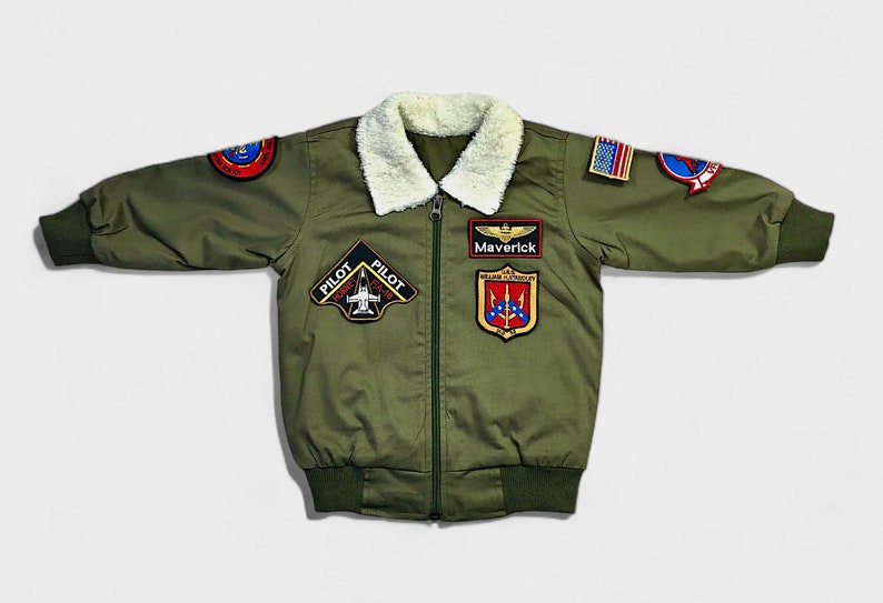 K&ouml;nnte beinhalten: Eine olivgr&uuml;ne Bomberjacke mit wei&szlig;em Kunstfellkragen, Frontrei&szlig;verschluss und gerippten B&uuml;ndchen. Die Jacke ist mit verschiedenen gestickten Aufn&auml;hern verziert, darunter ein Aufn&auml;her mit der Aufschrift Maverick, eine US-Flagge und eine Pilot F/A-18 Hornet Grafik.