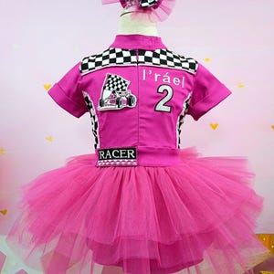 May include: Pink and black checkered racing-themed tutu dress with a pink tulle skirt and a black "RACER" patch. The dress has a pink and white bow on the back and a patch with a cartoon race car and the text "l'raël 2".