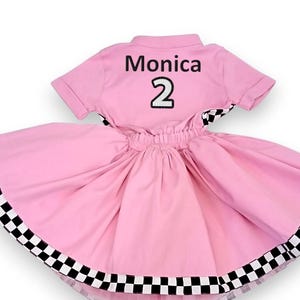 May include: Pink and black checkered dress with a white collar and short sleeves. The back of the dress has the name "Monica" and the number "2" embroidered in black.