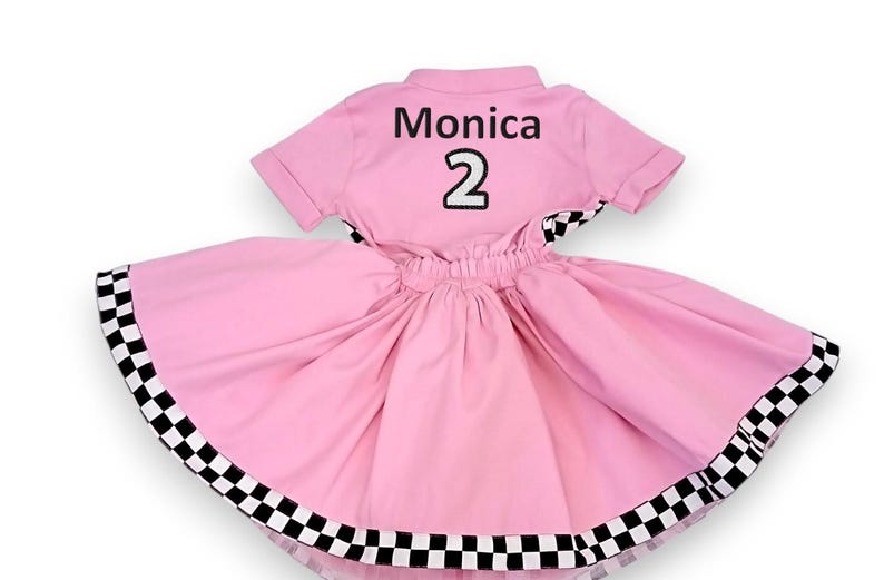 May include: Pink and black checkered dress with a white collar and short sleeves. The back of the dress has the name "Monica" and the number "2" embroidered in black.
