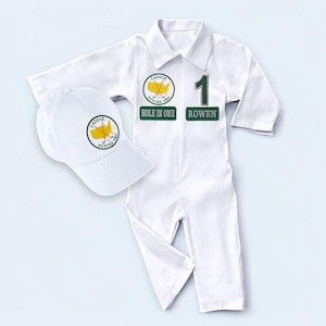 Front-facing shot of the white baby golf caddy romper. The garment features a prominent circular golf tournament logo on the left chest and a bold number 1 on the right. A matching white toddler hat is placed beside it, showcasing a complete, ready-to-wear set for young sports fans or as a unique birthday gift idea.
