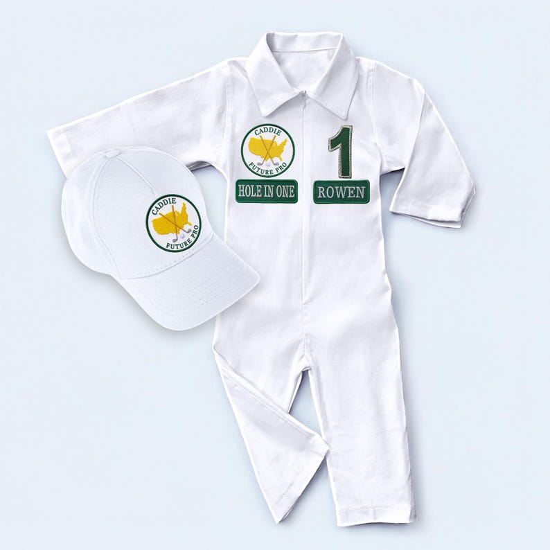 Front-facing shot of the white baby golf caddy romper. The garment features a prominent circular golf tournament logo on the left chest and a bold number 1 on the right. A matching white toddler hat is placed beside it, showcasing a complete, ready-to-wear set for young sports fans or as a unique birthday gift idea.