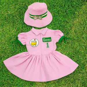 Op de afbeelding: Een roze jurk met korte mouwen en een witte kraag. De jurk heeft een groen lint op elke mouw en een groene patch met een gele golfbal en de tekst "Masters" aan de linkerkant van de borst. De jurk heeft ook een groene patch met de tekst "Emma 1" aan de rechterkant van de borst. Een roze hoed met een groen lint ligt bovenop de jurk.
