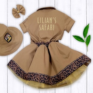 May include: A brown safari-themed dress with a leopard print trim and a matching hat. The dress has a white embroidered patch that says "Lillian's Safari".