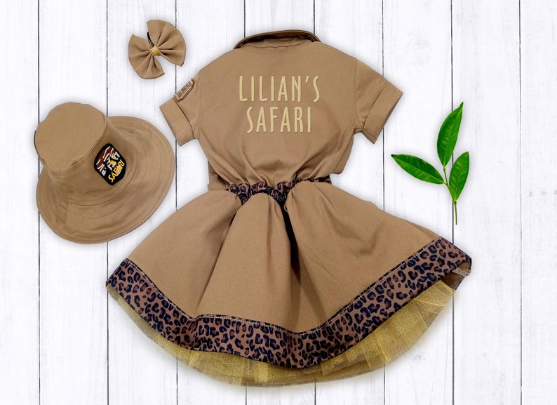 May include: A brown safari-themed dress with a leopard print trim and a matching hat. The dress has a white embroidered patch that says "Lillian's Safari".