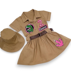 May include: A tan safari-style dress and matching hat. The dress has a green dinosaur patch, a pink number 2 patch, and a pink dinosaur footprint. The name "Eleanor" is on a patch. A brown belt defines the waist.