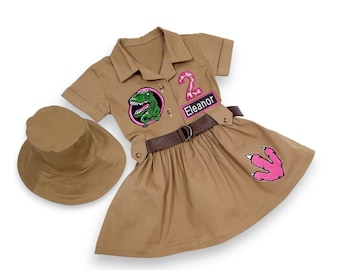 May include: Tan safari-style dress with a matching hat. The dress features a green dinosaur patch, a pink number 2 patch, and a pink dinosaur footprint. The name "Eleanor" is on a patch. A brown belt cinches the waist.
