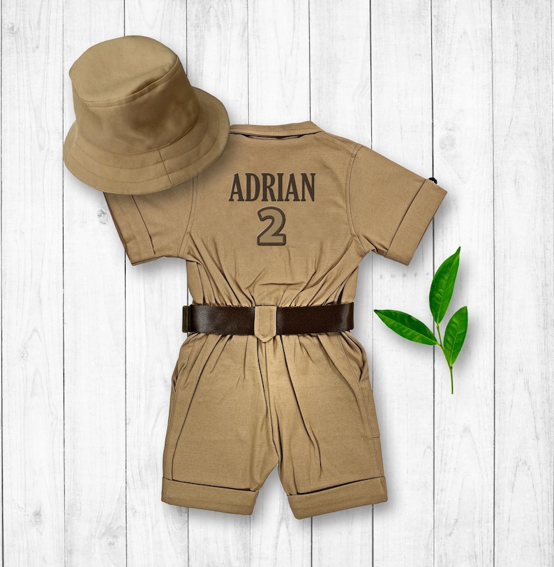 May include: A tan safari jumpsuit with a brown belt and a brown hat. The jumpsuit has the name "ADRIAN" and the number "2" printed on the back.