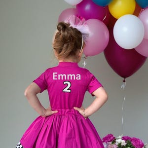 May include: A magenta dress with a black and white checkered hem, featuring the name "Emma" and the number "2" on the back. A collection of colorful balloons and a bouquet of pink flowers are in the background.