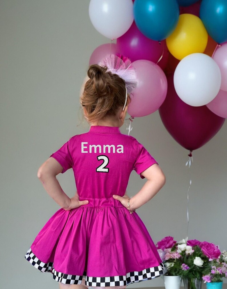 May include: A magenta dress with a black and white checkered hem, featuring the name "Emma" and the number "2" on the back. A collection of colorful balloons and a bouquet of pink flowers are in the background.