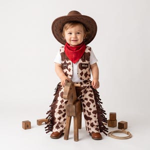 May include: A cowboy costume featuring a brown felt hat, a white t-shirt, a red bandana, and a vest and chaps set with a brown and white cow print pattern. The vest includes a sheriff badge and a name tag that reads Mick, with brown fringe.