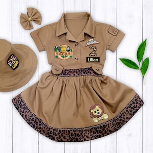 May include: A brown safari-themed dress with a lion patch, a leopard print trim, and a matching hat. The dress has a patch that says "Safari" and another patch with the number "2" and the name "Lillian".