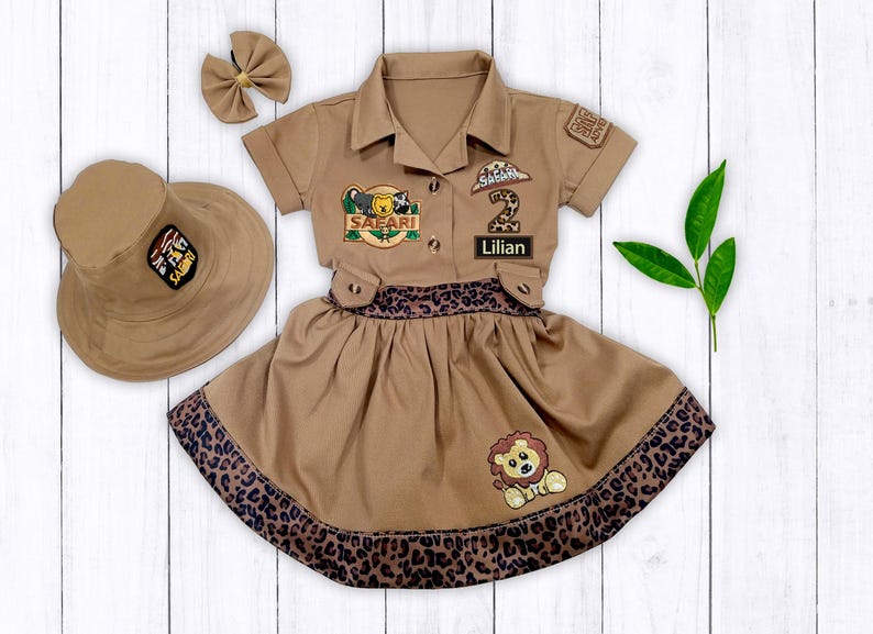 May include: A brown safari-themed dress with a lion patch, a leopard print trim, and a matching hat. The dress has a patch that says "Safari" and another patch with the number "2" and the name "Lillian".