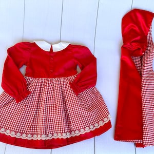 K&ouml;nnte beinhalten: Rotes und wei&szlig;es Gingham-Kleid mit wei&szlig;em Kragen und passender roter Kapuze. Das Kleid hat einen wei&szlig;en Spitzenbesatz.